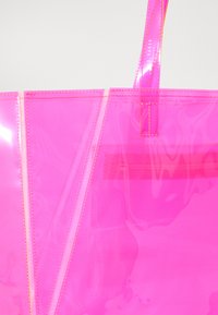 Transparente pinkfarbene Tragetasche mit glänzender Oberfläche, mit genähten Kanten und zwei Griffen. Enthält eine innere Reißverschlusstasche.
