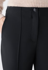 Pantalones negros confeccionados en tejido suave con frente plano, bolsillos laterales y un botón decorativo en la cintura.