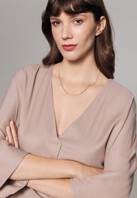 Beżowa bluzka z głębokim dekoltem w kształcie litery V i szerokimi rękawami; na szyi noszona jest delikatna złota łańcuszek. Gładka faktura, minimalistyczny design.