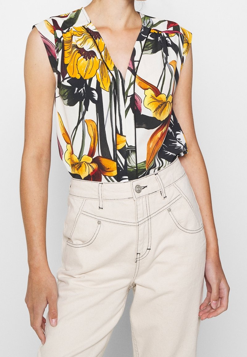 Blouse sans manches avec imprimé floral jaune, orange et noir, associée à un pantalon crème taille haute avec coutures visibles.
