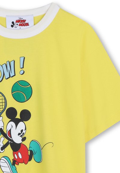 T-shirt jaune avec col blanc présentant Mickey Mouse jouant au tennis, frappant une balle de tennis verte avec une raquette et le texte "WOW!" au-dessus.