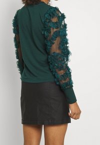 Top en teal con mangas de tela transparente adornadas con acentos de flores texturizadas en teal, combinado con una mini falda negra.