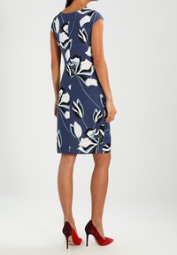 Robe bleu marine à fleurs avec manches courtes, présentant des motifs de fleurs blanches et bleues. Portée avec des chaussures à talons rouges. Tissu lisse, silhouette ajustée.