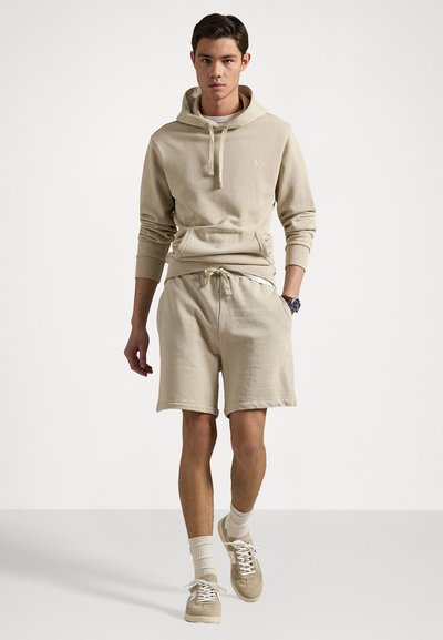 Bež hoodie s vezicom, kenguru džepom i odgovarajućim kratkim hlačama. Mekana tekstura, opušteni kroj, uparen s bež tenisicama i bijelim čarapama.