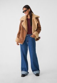 Braune Schaflederjacke mit breitem Kragen, kombiniert mit blauen weit geschnittenen Jeans. Eine Sonnenbrille und ein figurbetontes burgunderfarbenes Oberteil runden das Outfit ab.