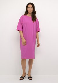 Przesadna sukienka t-shirt w jaskrawym różu. Wykonana z miękkiej bawełny, z okrągłym dekoltem, krótkimi rękawami, sięgająca tuż nad kolana. Noszona z czarnymi klapkami.