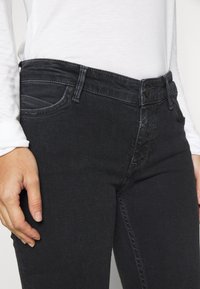 Jeans de mezclilla negra con cintura de altura media, diseño de cinco bolsillos y un sutil desgaste en la parte frontal. Textura suave, corte ajustado.