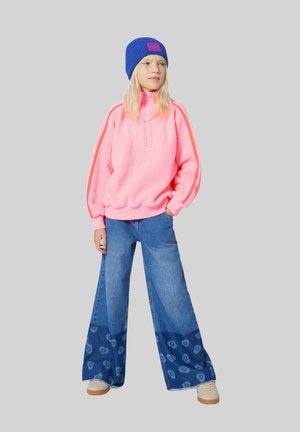 Fleece pullover in felroze met een kwart rits; wijde denim broek met een blauw-wit paisleypatroon; blauwe gebreide beanie.