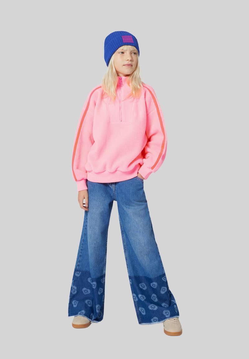 Pullover en polaire rose vif avec une fermeture éclair quart ; pantalons en denim à jambe large présentant un motif paisley bleu et blanc ; bonnet en tricot bleu.