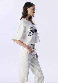 Weißes, kurzes T-Shirt mit schwarzem Grafikdesign, kombiniert mit hoch taillierten, weißen weit geschnittenen Hosen mit floraler Stickerei am Bein.