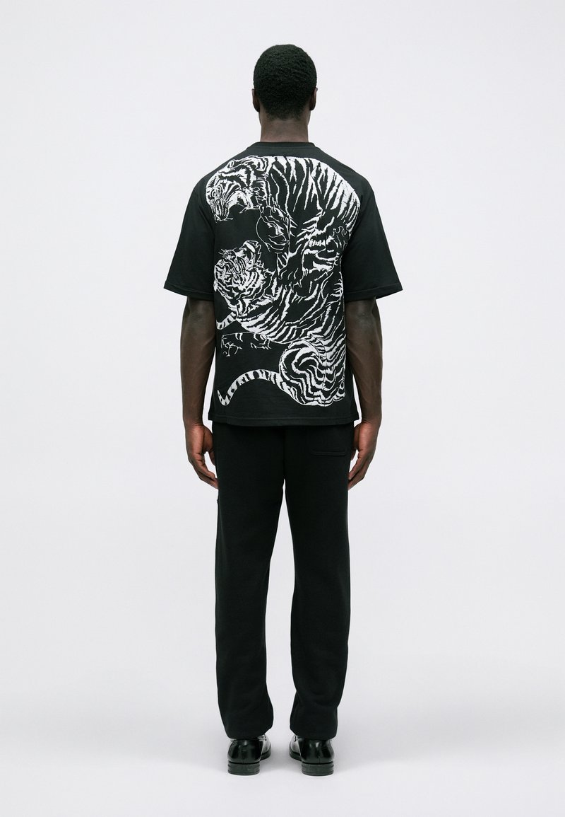 Homme debout de dos portant un t-shirt noir avec deux tigres blancs dans une pose dynamique au dos, un pantalon noir et des chaussures noires.