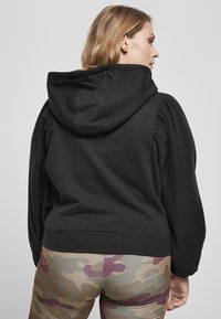 Felpa nera con maniche a palloncino, tessuto testurizzato e cappuccio ampio. Abbinata a leggings con motivo mimetico chiaro in tonalità terrose.