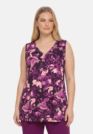 Vrouw met een mouwloze paarse top met roze bloemenprint en bijpassende paarse broek, staand tegen een effen achtergrond.