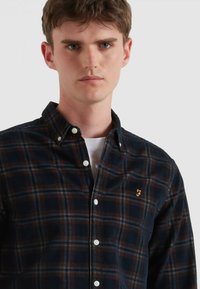 Camisa a cuadros en azul marino y marrón, hecha de una tela suave, con cuello abotonado y bordado de marca visible en el pecho.