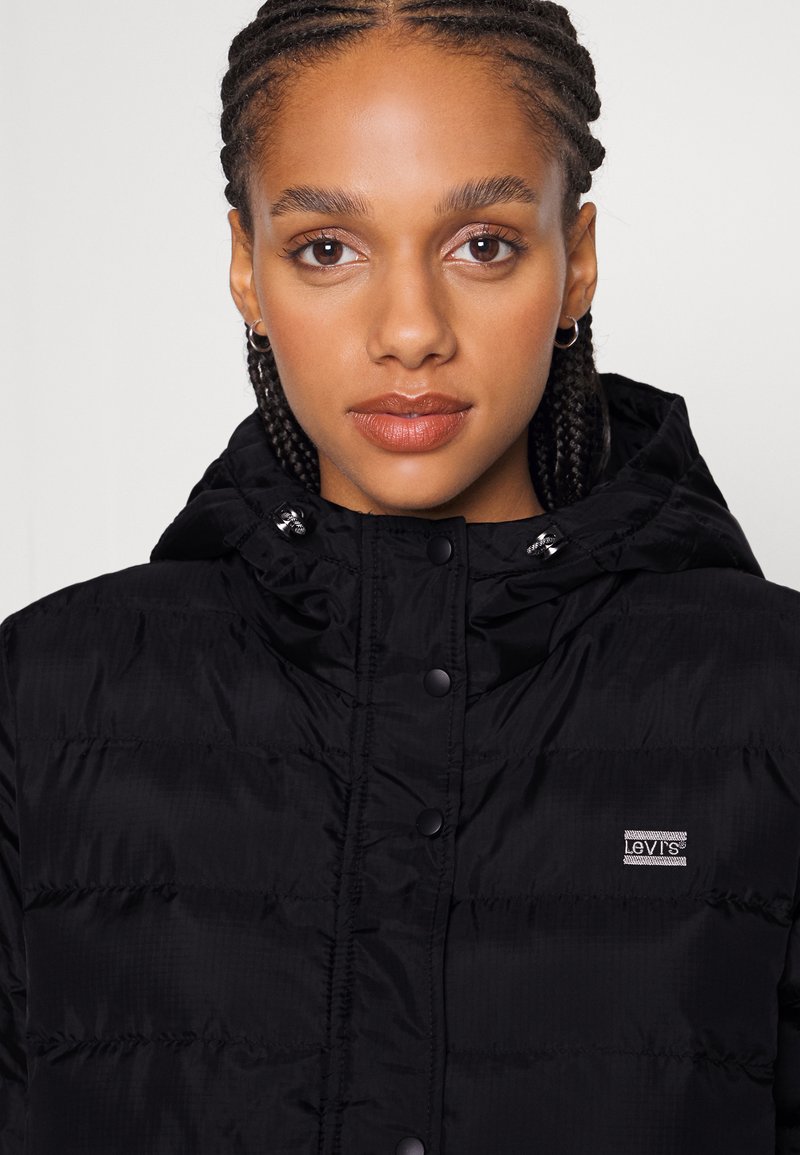 Doudoune noire avec capuche, présentant une texture brillante et lisse ainsi que des boutons-pression. Affiche un petit logo Levis sur le côté gauche.