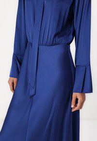 Abito in satin blu con vita legata, maniche lunghe e svasate, e una trama liscia. Presenta un design plissettato per un ulteriore profondità.
