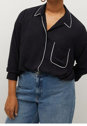 Personne portant une chemise noire boutonnée avec un passepoil blanc et une poche poitrine, associée à un jean bleu taille haute.