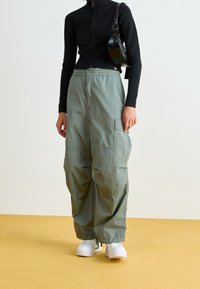 Person som bär en svart tröja med långa ärmar, lösa gröna cargobyxor med snörning vid bensluten, vita sneakers och bär en svart axelväska.