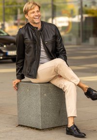 Black leather jacket with zip pockets, gray shirt, beige trousers, and black dress shoes, seated on a granite block outdoors.