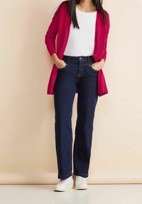 Červený cardigan přes bílé tričko, v kombinaci s tmavě modrými džíny rovného střihu a bílými teniskami. Džíny mají kapsy. Hladké textury.