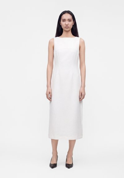 Femme portant une robe midi blanche sans manches et des talons noirs pointus, debout devant un fond blanc uni avec une expression neutre.