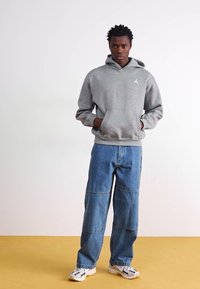 Sweat à capuche gris avec poche avant et logo, associé à un jean en denim bleu clair ample. Modèle portant des chaussures de sport blanches.