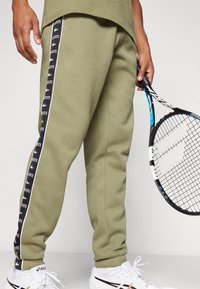 Pantalones de chándal verde oliva con rayas laterales en blanco y negro que presentan logotipos, con tobillos ajustados y acompañados de una raqueta de tenis.