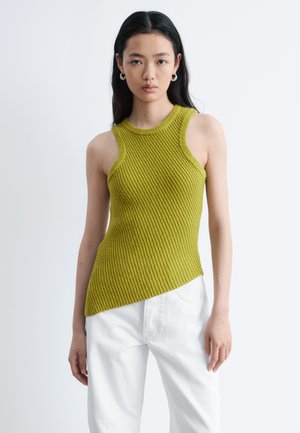 Jeune femme aux longs cheveux foncés portant un haut sans manches côtelé vert olive et un pantalon blanc, debout devant un fond clair uni.