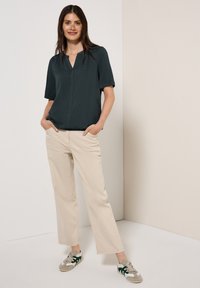 Blusa verde scuro a maniche corte con scollo a V, abbinata a pantaloni larghi beige chiaro e sneakers grigie con dettagli verdi.