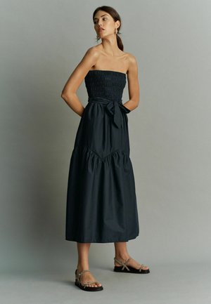 Svart axelbandslös maxiklänning med smockad bodice, knyt-dégradé i midjan, och lagerkjol. Modellen har på sig beige sandaler, mot en neutral bakgrund.