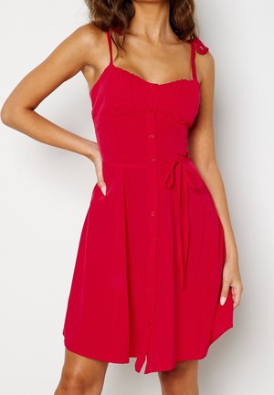 Mujer con vestido rojo sin mangas con botones en la parte delantera, tirantes finos y cinturón anudado en la cintura, mano en el bolsillo, sobre un fondo liso.