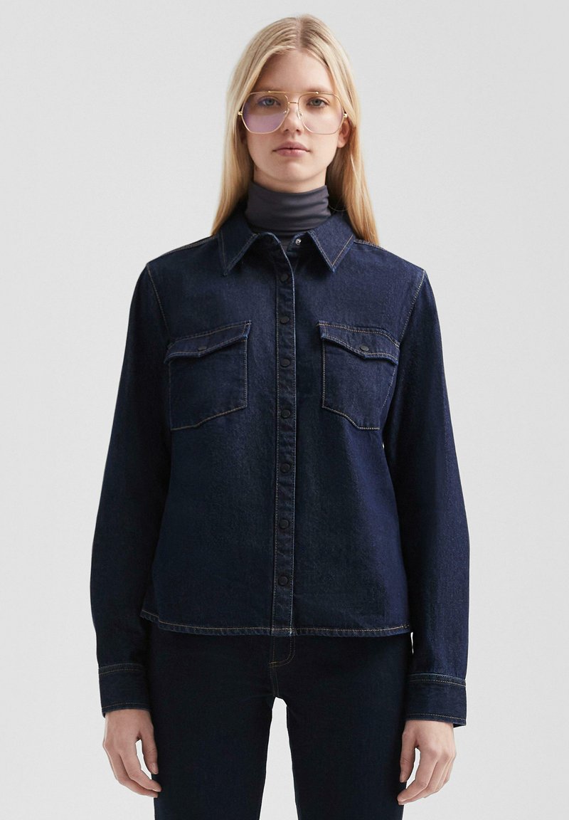 Giubbotto in denim blu scuro con colletto, due tasche sul petto, chiusura con bottoni e dettagli di cucitura visibili. Maniche lunghe, vestibilità relaxed.