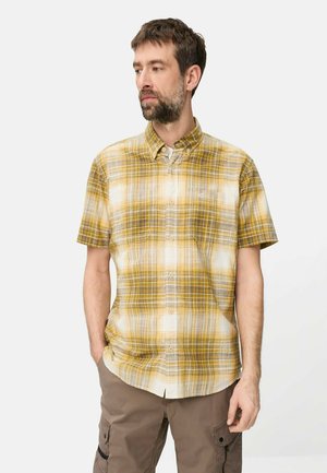 Mand med skæg iført en gul- og hvidternet kortærmet skjorte med knapper og brune cargobukser, stående med den ene hånd i lommen.