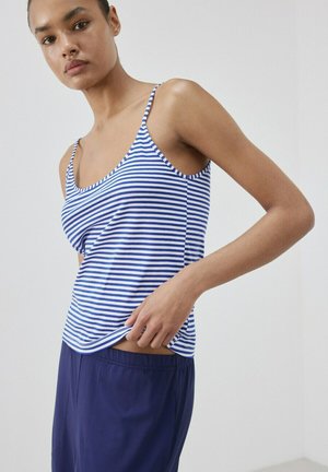 Femme ajustant un débardeur rayé bleu et blanc, portant un pantalon ample bleu marine, sur un fond clair uni.