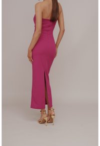 Abito rosa aderente senza spalline con uno spacco laterale e una texture liscia. Indossato con sandali oro con tacco alto. La vista posteriore mette in risalto la silhouette.