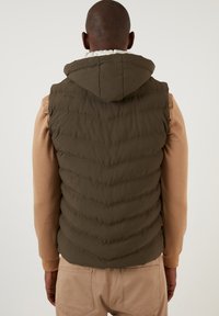 Olive green padded vest with hood, featuring chevron quilted design. Worn over light brown long-sleeve shirt, with visible stitching detail.