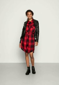 Vestido camponês em padrão escocês vermelho e preto com botões, cintura ajustada, mangas curtas, usado sob uma jaqueta de couro preta e combinado com botins pretos.
