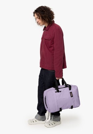 Sac en tissu violet avec des poignées noires, de forme rectangulaire et fermeture éclair. Porté par une personne en veste bordeaux et jeans foncés.