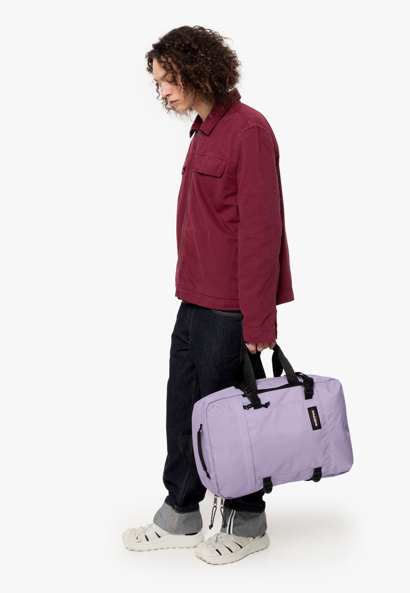 Lila tygväska med svarta handtag, rektangulär form och dragkedjestängning. Bärs av en person i en vinröd jacka och mörka jeans.