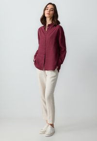 Camicia di lino bordeaux con frontale a bottoni, maniche lunghe e tasche laterali, abbinata a pantaloni di lino beige chiaro e sneaker bianche.