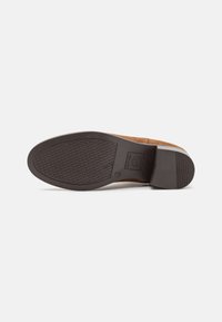 Semelle d'une chaussure en daim marron avec une semelle en caoutchouc texturée, un logo rectangulaire près de la voûte plantaire et un talon bloc, sur fond blanc.