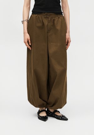 Kvinde iført løse brune ballonbukser med elastiske manchetter, en sort top, sølvarmbånd og sorte spidse flade sko med nitter.