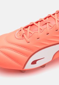 Crampons de football synthétiques orange avec un dessus texturé, des accents cousus, un logo blanc proéminent et une semelle à multiples crampons pour une meilleure adhérence.