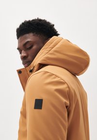 Chaquetón resistente al agua de color mostaza con una gran capucha, cierre de cremallera y a presión, y un parche negro con el logo en la manga izquierda.