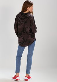 Langermet hettegenser i mørkt camo-mønster, laget av mykt stoff. Kombinert med blå skinny jeans og røde joggesko.