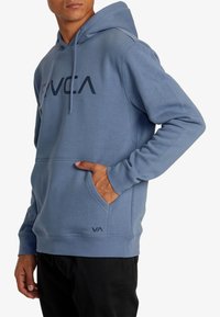 Felpa blu in pile con un grande logo sul davanti, cappuccio con cordoncino, tasca a marsupio e polsini a coste. Logo discreto sull'orlo.
