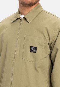 Tan werkjack met een ritsluiting aan de voorkant, puntige kraag en een borstzak met een zwart logo-patch. Gemaakt van stevig materiaal.