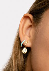 Goldene Creolen mit einem kleinen lächelnden Gesichtchen-Anhänger und einem hellblauen Akzentring, präsentiert vor einem neutralen Hintergrund.