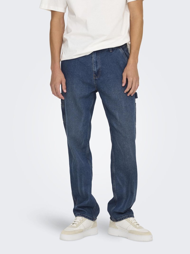 Man draagt een blauwe rechte spijkerbroek, witte sneakers en een wit shirt met korte mouwen en staat voor een effen achtergrond.