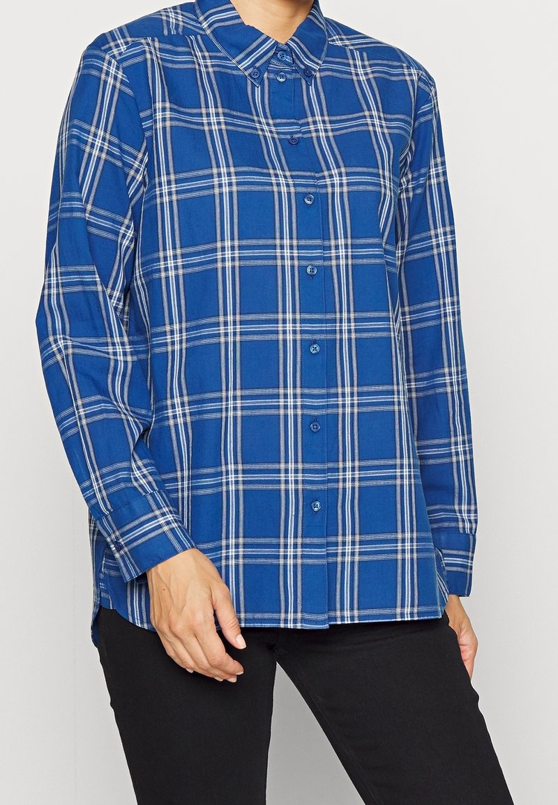Camisa de cuadros azul de botones, hecha de algodón, con mangas largas, cuello y un patrón a cuadros con acentos en blanco y azul más oscuro.
